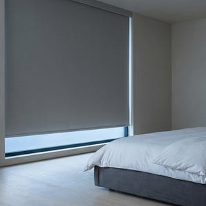 May include: A gray roller shade covers a window in a bedroom. The shade is pulled down, blocking most of the natural light. A bed with white bedding is in the foreground. The room has a minimalist design with neutral colors.