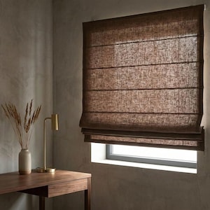 May include: A dark brown Roman shade made of a textured fabric with horizontal seams, installed over a window. The shade features a woven appearance and is partially lowered, allowing light to filter through the material into the room.