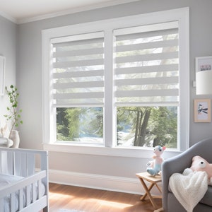 K&ouml;nnte beinhalten: Wei&szlig;e Fensterjalousien mit horizontalem Streifenmuster, die zwei Fenster in einem Raum mit wei&szlig;en W&auml;nden und einem Holzboden bedecken. Ein wei&szlig;es Kinderbett und ein grauer Sessel mit einem Stofftier sind im Raum zu sehen.