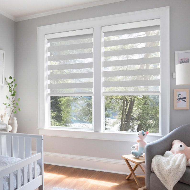 K&ouml;nnte beinhalten: Wei&szlig;e Fensterjalousien mit horizontalem Streifenmuster, die zwei Fenster in einem Raum mit wei&szlig;en W&auml;nden und einem Holzboden bedecken. Ein wei&szlig;es Kinderbett und ein grauer Sessel mit einem Stofftier sind im Raum zu sehen.
