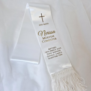 May include: A white satin baptism sash with gold text and a gold cross. The sash includes the text "GOD BLESS", a name, and baptism details: date, church, and location. The bottom edge has a white fringe.
