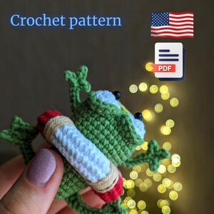 May include: Crochet pattern for a green frog wearing a red and white life preserver. The pattern is available as a PDF download. The image shows the finished crochet frog.