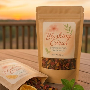 May include: Two kraft paper pouches of "Blushing Citrus" simmer blend fragrance. One pouch is open, spilling the blend and a slice of orange. The label features a floral design and reads "Simmer Blend Fragrance" and "Net Wt. 1.2 oz."