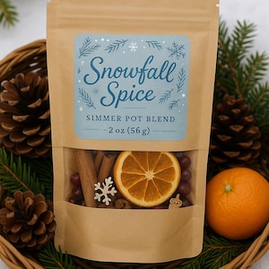 May include: A brown paper bag with a clear window, labeled "Snowfall Spice Simmer Pot Blend - 2 oz (56 g)". The bag contains cinnamon sticks, dried orange slices, and other spices. The bag is in a wicker basket with pine cones and an orange.
