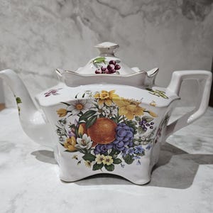 May include: A white ceramic teapot with a scalloped rim and a matching lid. The body features a colourful botanical illustration of yellow daffodils, purple grapes, and a central orange fruit, with additional small floral accents on the spout and lid.