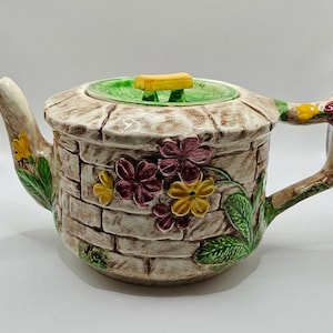 May include: A ceramic teapot featuring a textured brick wall pattern in shades of beige and brown. The exterior is decorated with raised, colourful floral motifs in purple, yellow, and green. It includes a matching lid with a yellow handle.