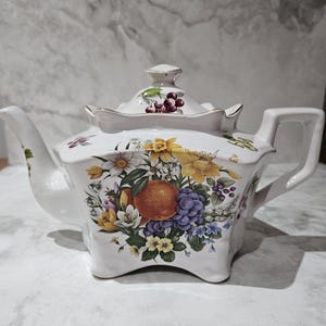 May include: White ceramic teapot with a square base and a lid. The teapot is decorated with a colorful floral and fruit design, including oranges, grapes, and various flowers. The handle and spout are white.