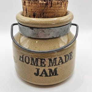 May include: A tan ceramic jar with a speckled finish, featuring a metal handle and a cork stopper. The text "HOME MADE JAM" is printed in black on the front. The jar has a rounded body and a thick rim, providing a sturdy container for storage.
