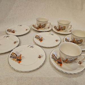 以下が含まれることがあります： 航海をテーマにしたヴィンテージのティーカップとソーサーのセット6個。カップとソーサーは、白い背景にカラフルな帆船と海辺の町のイラストが描かれています。