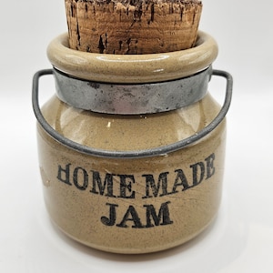 May include: A small, tan ceramic jar with a cork lid and a wire handle. The jar has the words "HOME MADE JAM" printed on the front.