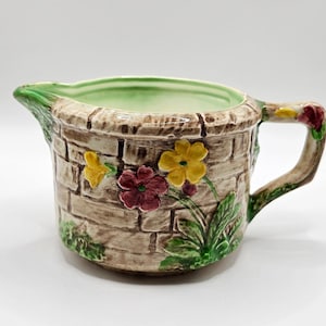 May include: A ceramic jug with a brick-effect exterior, decorated with yellow and maroon flowers and green leaves. The handle and rim have floral details. The interior is a pale green colour.