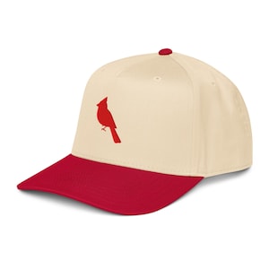 May include: A baseball cap with a cream-colored crown and a red brim. A red cardinal bird silhouette is on the front of the cap. The cap has a curved brim and a button on top.
