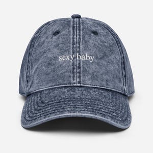 May include: A denim-style baseball cap in a faded blue color. The cap features the embroidered text "sexy baby" in white, centered on the front. The cap has a curved brim and an adjustable strap.