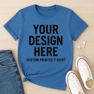 May include: A blue t-shirt with the text "YOUR DESIGN HERE" in large black letters. Below the text is "CUSTOM PRINTED T-SHIRT". The shirt is paired with blue jeans and white sneakers.