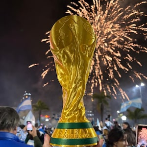 May include: A gold-coloured replica of the FIFA World Cup trophy featuring a globe supported by two figures, with a green base displaying the text FIFA WORLD CUP in gold lettering, held against a night sky filled with bright orange fireworks.