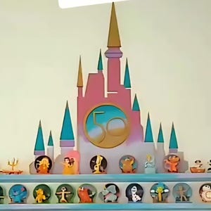 Puede incluir: Una estantería decorativa con un castillo rosa y dorado con el número 50. La estantería exhibe varias figuras de personajes de Disney, incluyendo a Ariel, Mickey Mouse y Stitch, dispuestas en dos niveles.