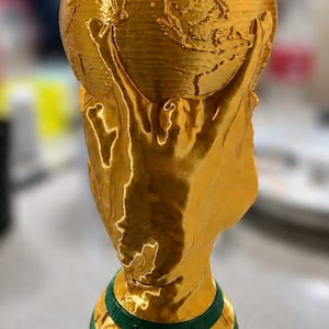 May include: A gold-coloured trophy cup featuring a raised world map design and two figures with arms raised. The base is green with the text "ARGENTINA CAMPEON" embossed in gold. A silver-coloured rim tops the cup, and a black label reads "@laserwech".