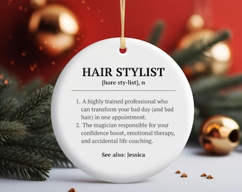 May include: A round white ceramic ornament featuring black text that defines a hair stylist as a professional who transforms bad hair and provides emotional therapy. The text includes the name Jessica and hangs from a gold string.