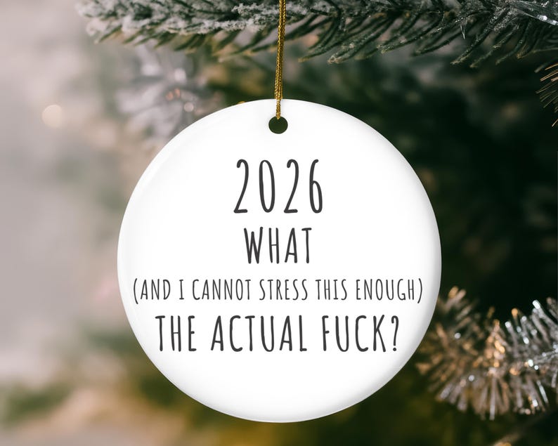 May include: White ceramic Christmas ornament with the text "2026 WHAT (AND I CANNOT STRESS THIS ENOUGH) THE ACTUAL FUCK?" in black. The ornament hangs from a gold string against a blurred background of a Christmas tree.
