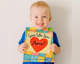 May include: A smiling child holds a colorful book titled "Guess Who Loves Marcus?" The book cover features a red heart and the name "Marcus." The book has a patchwork design with various colors.