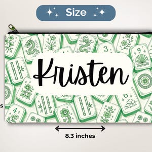 May include: A rectangular zippered pouch featuring a green and white pattern of mahjong tiles with various symbols, including dragons, bamboo, and flowers. The center displays the name Kristen in black script. It measures 4.7 inches by 8.3 inches.