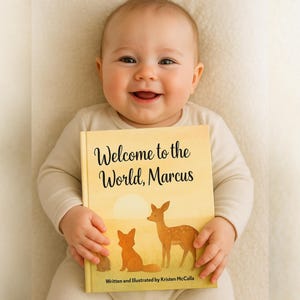 May include: A smiling baby holds a book titled "Welcome to the World, Marcus." The book cover features illustrations of a fox and a deer in a yellow landscape. The text "Written and Illustrated by Kristen McCalla" is at the bottom.