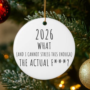 May include: A white ceramic Christmas ornament with the text "2026 WHAT (AND I CANNOT STRESS THIS ENOUGH) THE ACTUAL F***?" in black. The ornament hangs from a gold string and is surrounded by a Christmas tree.
