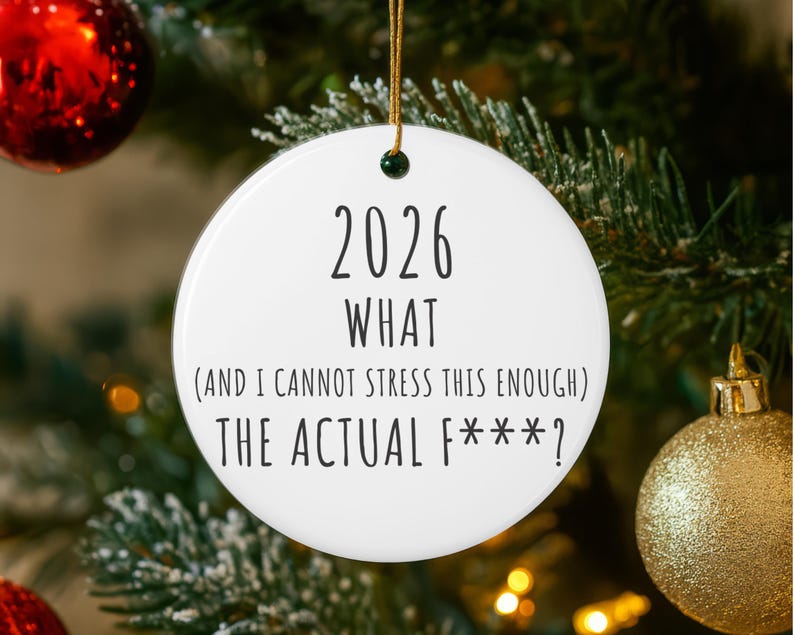 May include: A white ceramic Christmas ornament with the text "2026 WHAT (AND I CANNOT STRESS THIS ENOUGH) THE ACTUAL F***?" in black. The ornament hangs from a gold string and is surrounded by a Christmas tree.