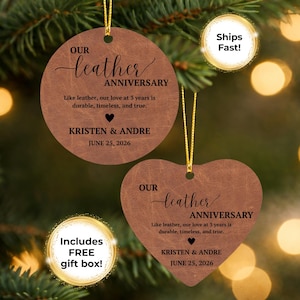 May include: Two brown leather-textured ornaments, one circular and one heart-shaped, featuring black printed text: OUR leather ANNIVERSARY, Like leather, our love at 3 years is durable, timeless, and true, KRISTEN & ANDRE, and JUNE 25, 2026.