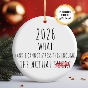 May include: A white ceramic Christmas ornament with the text "2026 WHAT (AND I CANNOT STRESS THIS ENOUGH) THE ACTUAL F***?" in black. A gold ribbon is attached for hanging. A gold circle with the text "Includes FREE gift box!" is in the upper right.