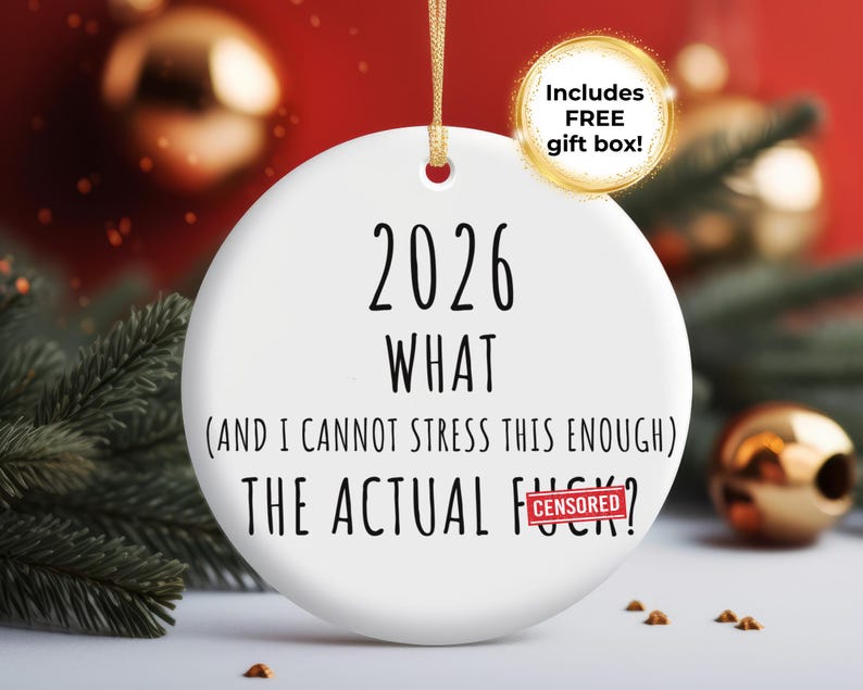 May include: A white ceramic Christmas ornament with the text "2026 WHAT (AND I CANNOT STRESS THIS ENOUGH) THE ACTUAL F***?" in black. A gold ribbon is attached for hanging. A gold circle with the text "Includes FREE gift box!" is in the upper right.