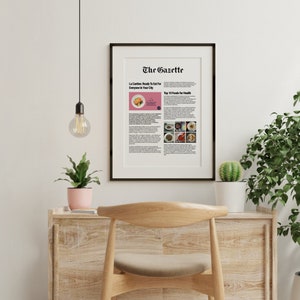 May include: A framed newspaper article titled "The Gazette" with the headline "La Cantine: Ready To Eat For Everyone In Your City" and a smaller headline "Top 10 Foods for Health". The article is surrounded by a white border and a black frame. The article is displayed on a wooden desk with a light bulb hanging from the ceiling above it.
