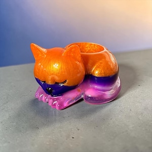 May include: A small, sleeping cat-shaped figurine with a hollow opening on its back. The head is a shimmering orange, the midsection is a deep purple, and the base and paws are a translucent pink. The cat has closed eyes and a small black nose.
