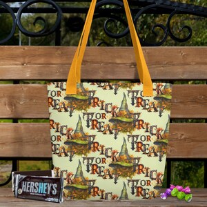 Puede incluir: Un bolso tote con temática de Halloween con un patrón repetido de sombreros de bruja y las palabras "Trick or Treat". El bolso tiene asas amarillas y se sostiene contra un banco de madera. Una barra de chocolate Hershey's y caramelos están en primer plano.
