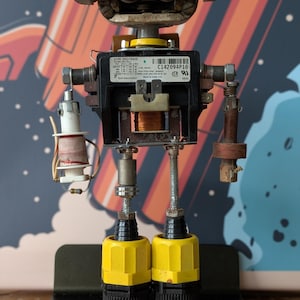 May include: A white ceramic mug with a black handle and the text "But First, Coffee" in a bold, black font. The mug is set against a blurred background, suggesting a cosy setting.