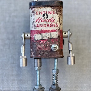 May include: A handcrafted robot sculpture made from recycled metal and a vintage "Handy Bandages" tin. The robot has a rusty, aged look with bolt and spring legs, and metal arm attachments. The robot is standing on a white surface.