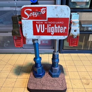 Könnte beinhalten: Eine Roboterskulptur aus recycelten Materialien. Der Kopf ist eine verwitterte weiße Metallbox mit rotem "Scripto"- und "VU-lighter"-Text. Rote und silberne Metallarme und -beine sind an einer Holzbasis mit einem topografischen Kartenmuster befestigt. Blaue und silberne Hardware wird verwendet.