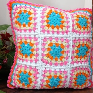 May include: A crocheted cushion with a white background and colourful squares. The squares are made of pink, orange, yellow, and blue yarn. The cushion has a scalloped edge.
