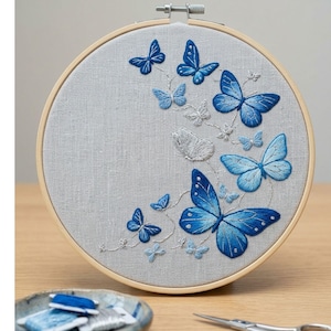 May include: A circular wooden embroidery hoop containing light gray fabric with a hand-stitched design of multiple butterflies in varying shades of blue and white. The butterflies are arranged in a flowing pattern across the right side of the hoop.