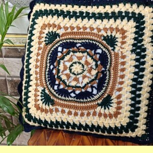May include: A square crocheted pillow with a blue, green, brown, and cream color pattern. The pillow has a scalloped edge and a detailed mandala design in the center.