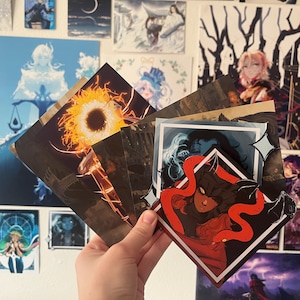 May include: A collection of five different illustrated cards featuring a variety of characters and designs. The cards are printed on a white background and have a glossy finish. The cards are held in a hand.