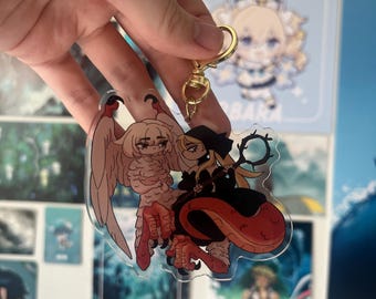 May include: A clear acrylic keychain featuring a cartoon illustration of a woman with long blonde hair wearing a black dress and a witch's hat, riding a red and white griffin with large wings.