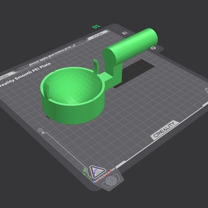 May include: A green 3D printed object with a bowl-shaped base and a curved handle. The object is sitting on a black and white grid surface. The text "Creality Smooth PEI Plate" is visible on the surface.