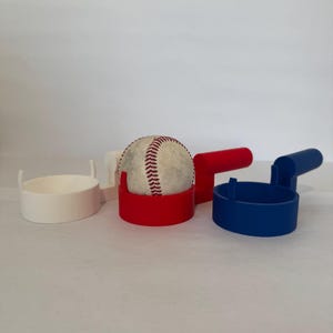 May include: Three baseball holders, one white, one red and one blue, with a baseball in the red holder.