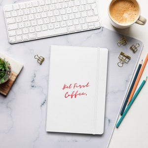 Pode incluir: Um caderno branco com uma inscrição vermelha que diz "But First Coffee." O caderno está sobre uma superfície de mármore branco com uma xícara de café, lápis e clipes.