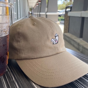 May include: A tan baseball cap with a small white embroidered design of a cartoon character holding a can. The character has a pink nose and black eyes.