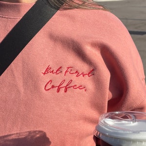 Könnte beinhalten: Ein rosa Sweatshirt mit dem Text "but first, coffee" in roter Stickerei. Das Sweatshirt wird von einer Person getragen, die eine Tasse Kaffee hält.