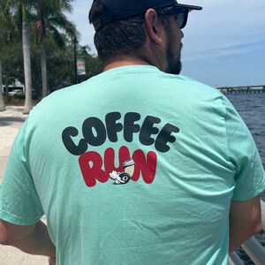 Pode incluir: Uma camiseta azul turquesa com o texto "COFFEE RUN" em letras pretas e vermelhas. Abaixo do texto, é retratado um copo de café de desenho animado com rodas.