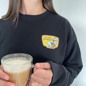 Pode incluir: Uma camisa preta de manga comprida com um gráfico amarelo e vermelho de uma xícara de café e o texto "All Caffe, No Brakes".