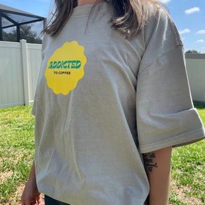 Pode incluir: Uma camiseta cinza claro com um gráfico amarelo e verde que diz "Addicted to Coffee".